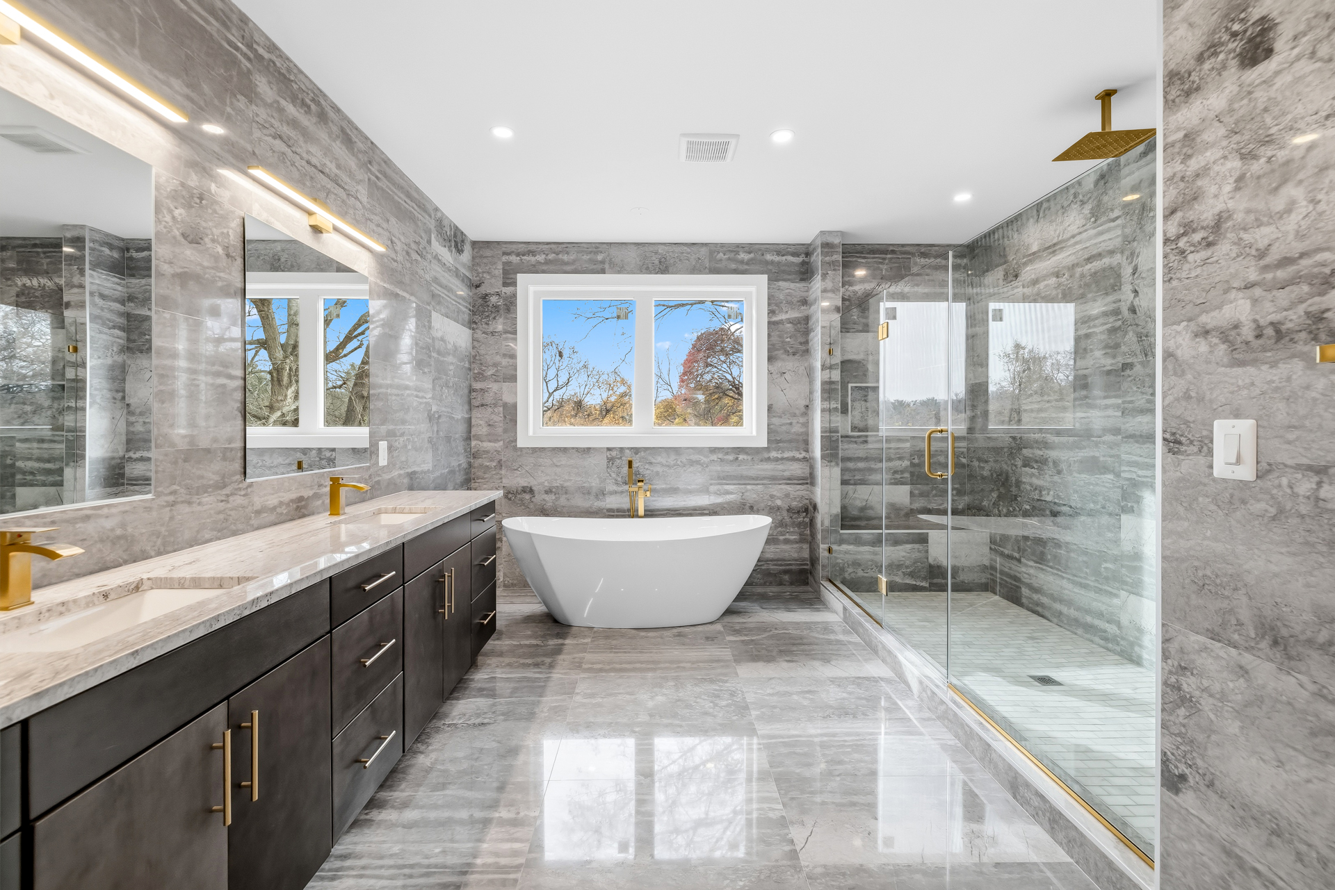 A luxury primary bathroom with gray marble tile, a freestanding soaking tub, a glass walk-in shower, and gold fixtures