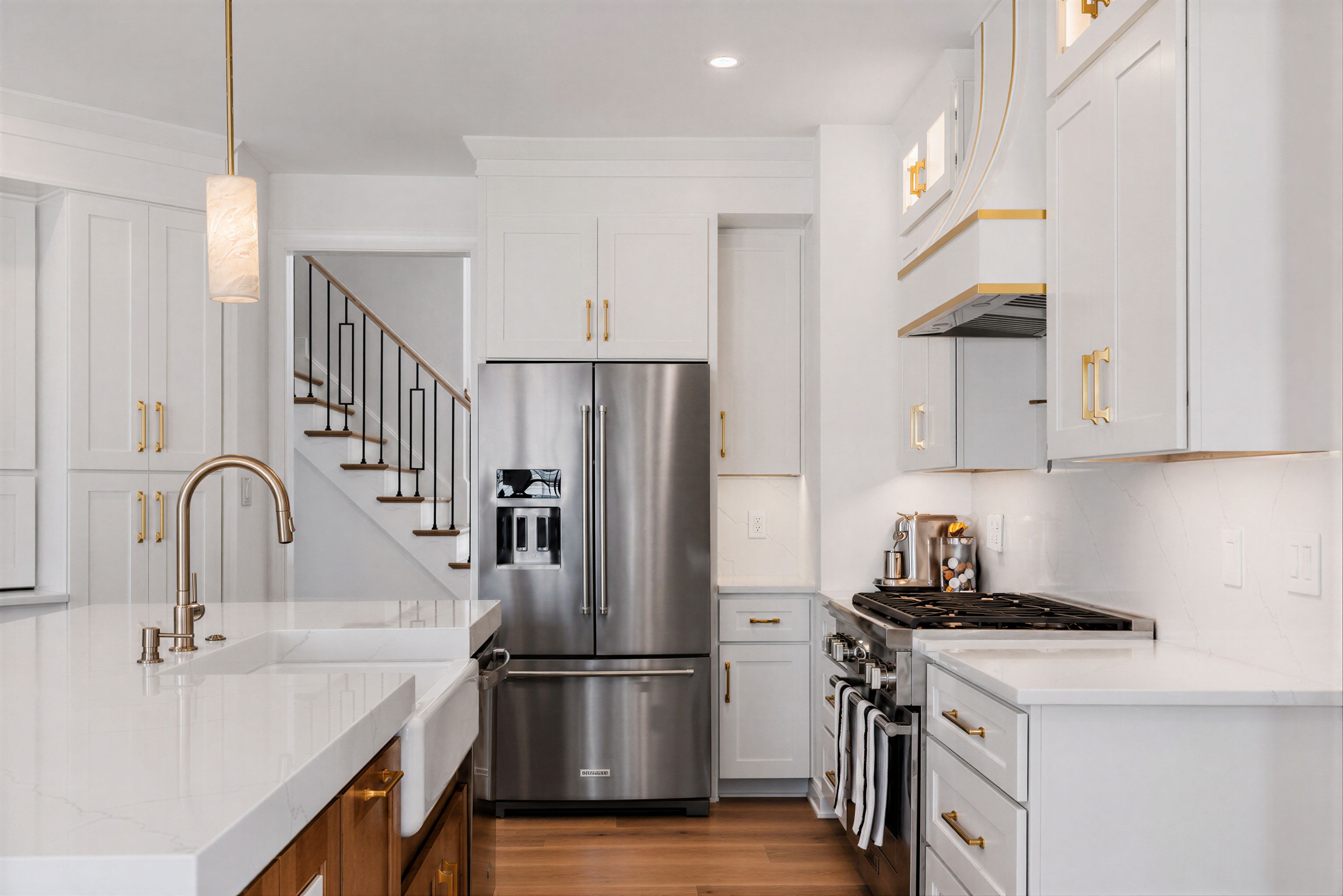 White kitchen with a farmhouse apron sink, brass hardware, a quartz island, and a staircase visible beyond